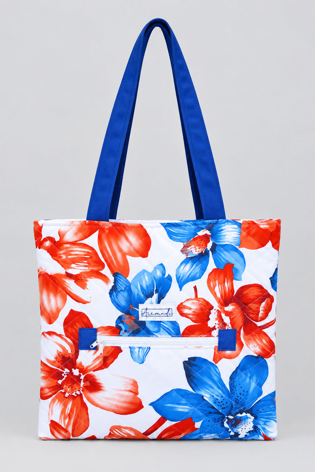 A white tote bag with a vibrant red and blue floral print, featuring two blue shoulder straps and a white zipper pocket in the front.