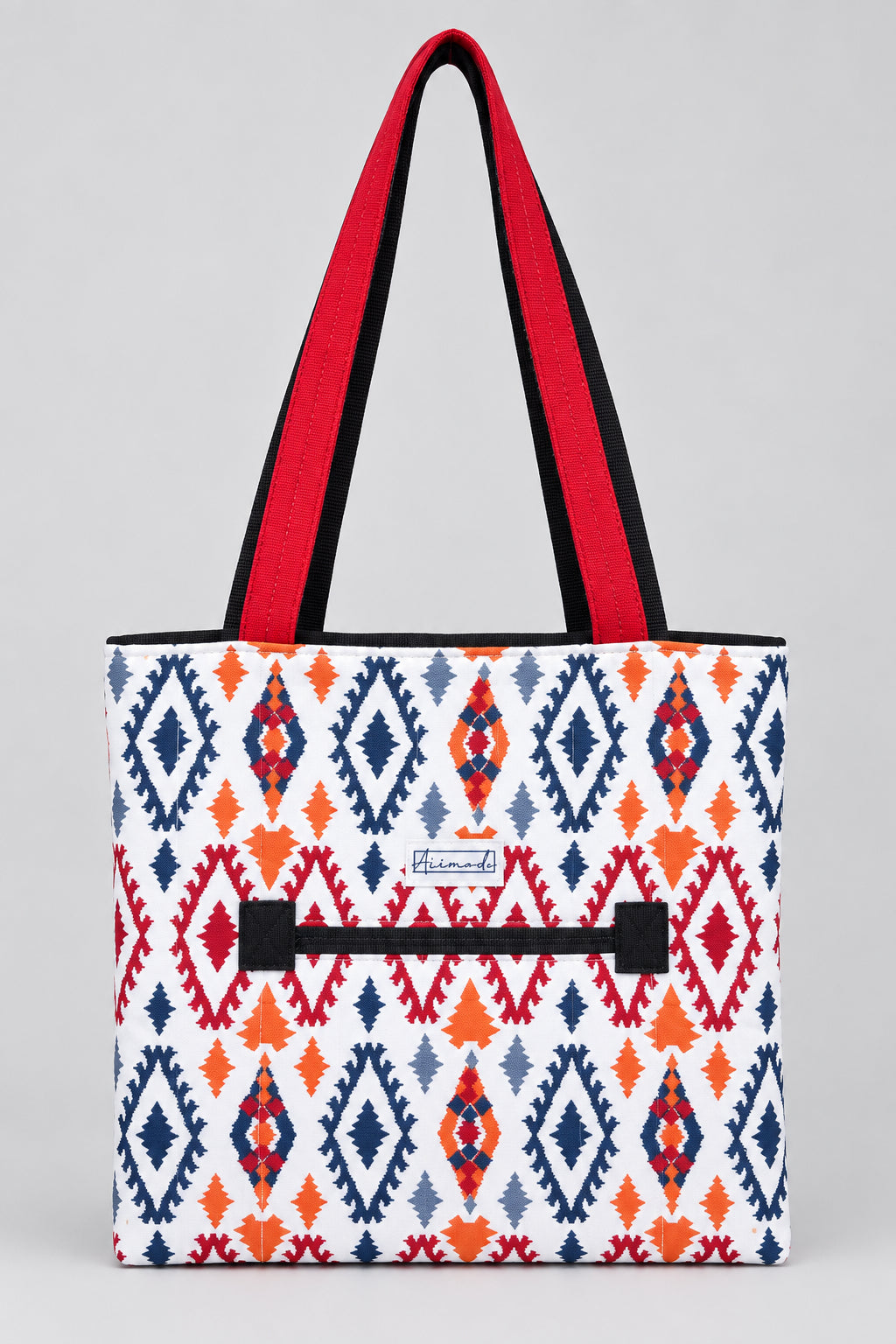 A tote bag with a white base featuring a colorful geometric diamond pattern in navy blue, red, and orange. It has bright red shoulder straps and a black zipper in the front.