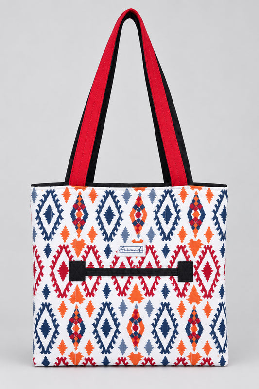 A tote bag with a white base featuring a colorful geometric diamond pattern in navy blue, red, and orange. It has bright red shoulder straps and a black zipper in the front.